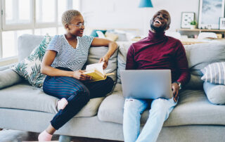 Couple sitting and laughing on couch, woman with book and man on laptop