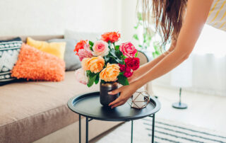 Woman putting small vase with colorful flowers on coffee table in living room